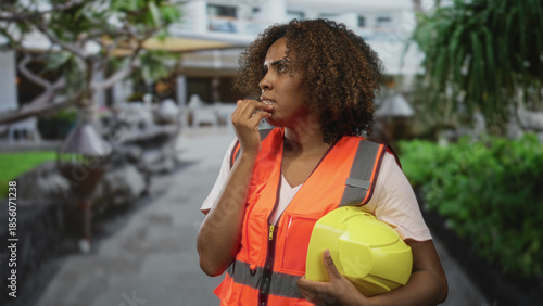 Foto Woman engineer wearing orange safety vest bites nails while holding yellow hardhat on city street near building; anxiety uncertainty