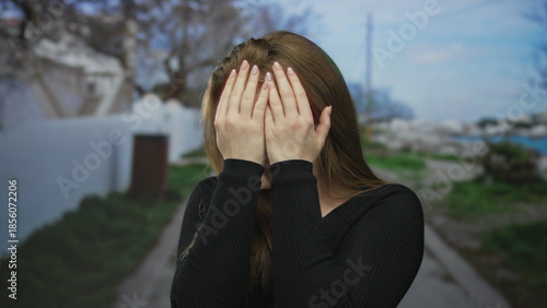 Schilderij op canvas Young caucasian woman in black sweater covers face with hands on seaside street; social embarrassment