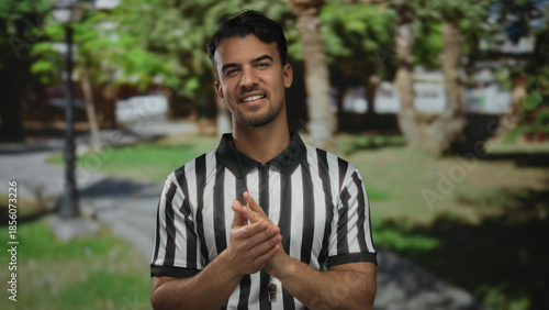 Young hispanic man clapping in a park wearing referee uniform outdoor with green trees and sunlight in the background showing happiness and sportsmanship