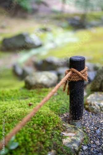 A thick rope is tied around a dark wooden post, marking a path through a mossy garden with scattered rocks.