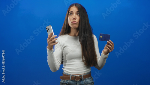 Photos Woman holding smartphone and creditcard, shrugs and lifts hands while making a puzzled face against blue studio backdrop; payment uncertainty