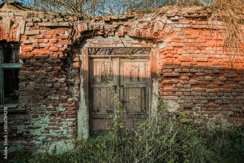 Old abandoned rural buildings. Decaying wooden door set in a cracked brick wall overgrown with plants. A symbol of passing time, neglect and forgotten places