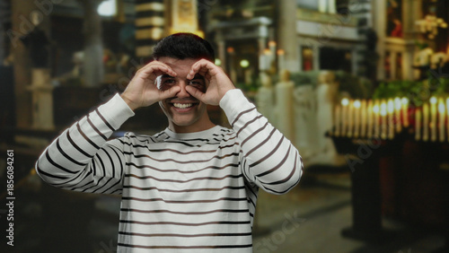 Obraz na plátně Young man in striped shirt mimicking binoculars indoors at church with candles in the background, conveying a playful and engaging atmosphere