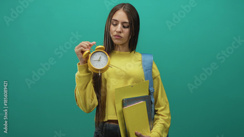 Foto Young woman student holding yellow alarm clock and folders, presenting time with raised hand in studio; deadline urgency impatience