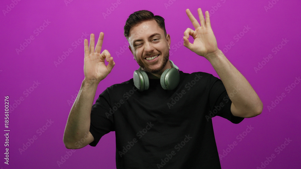Fototapeta premium Young man with headphones smiling and making an ok hand gesture against a vibrant pink background, exuding confidence and positivity.