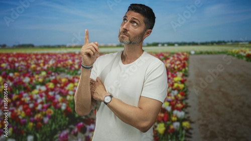 Man points finger upward with wristwatch visible and index finger raised in colorful tulip field studio near dirt path; contemplation.
