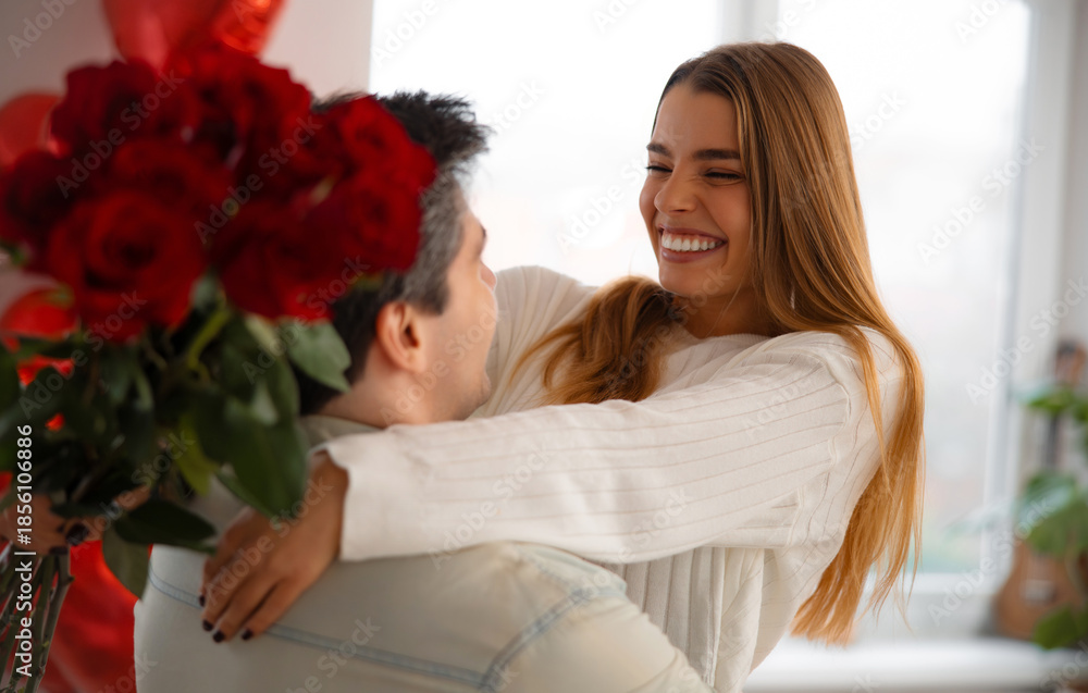 Obraz premium Smiling young woman hugging her partner while receiving a bouquet of red roses during a romantic Valentine’s Day moment at home. 