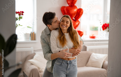 Young couple hugging and kissing at home with heart balloons, celebrating love and Valentine’s Day.
