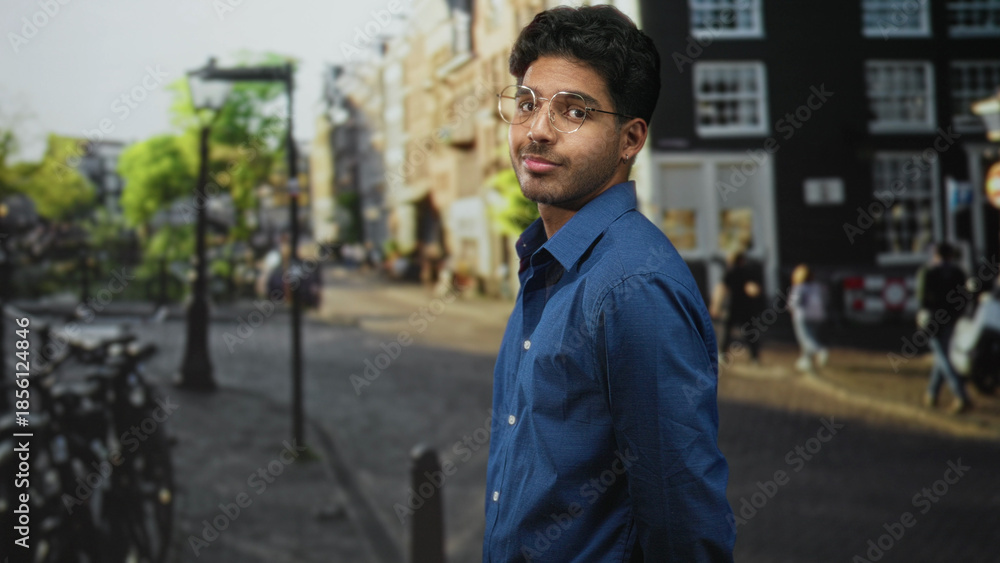 Fototapeta premium Young hispanic man wearing glasses and a blue shirt looks to the side showing his face on a city street; contemplation.