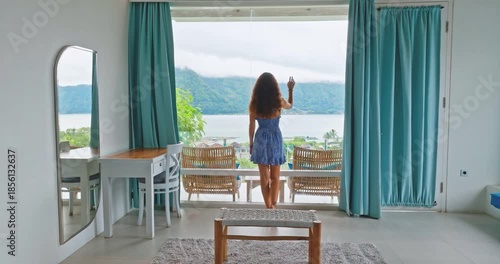Beautiful woman in short dress standing in hotel room, looking in full-length window balcony at serene Batur lake and mountains, summer travel and relaxation in Indonesia, Bali. Slow motion, back view