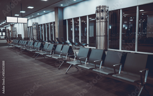Empty airport terminal waiting area at night with long row of metal seats, glass windows, ceiling lights, columns and information monitor, quiet departure gate interior; wide-angle, low-key shot