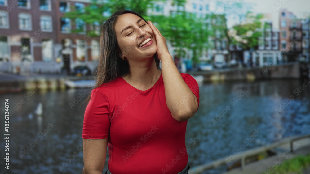 Fototapeta premium Woman in red shirt hand on cheek smiling on street by a canal with brick buildings in amsterdam; joy travel moment.