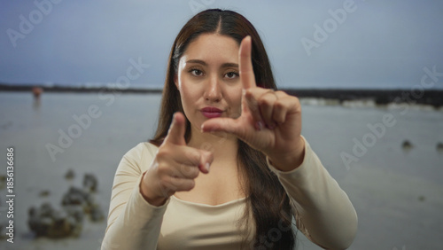 Woman holds a loser sign with her hand toward the camera on a windy beach with scattered rocks under an overcast sky at low tide; mockery.