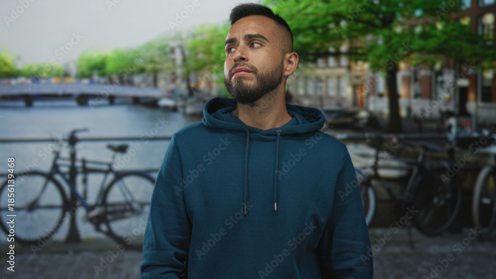 Fototapeta premium Man with hands on cheeks for toothache on amsterdam canal street near bicycles and bridge, wearing hoodie, looking pained; urban worry.