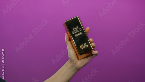 Obraz na plátně Man holding a fake gold bar against a vivid purple background, showcasing wealth concept in an isolated setting with focus on the hand's firm grip and reflective surface