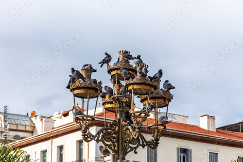A flock of pigeons perched on an ornate street lamp against a cloudy sky.