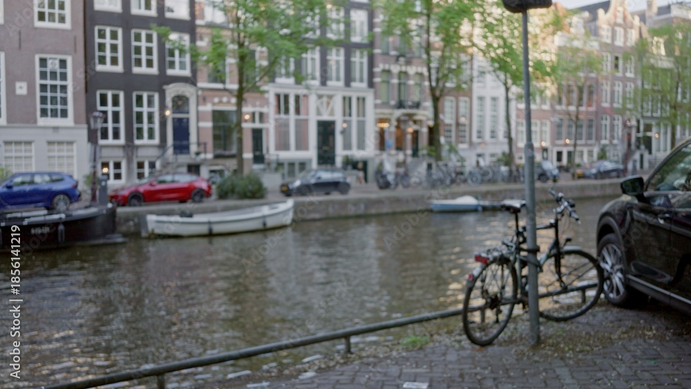 Fototapeta premium Bikes parked along the picturesque amsterdam canal under a blurred, defocused background of traditional dutch houses and vibrant trees reflecting in the water.
