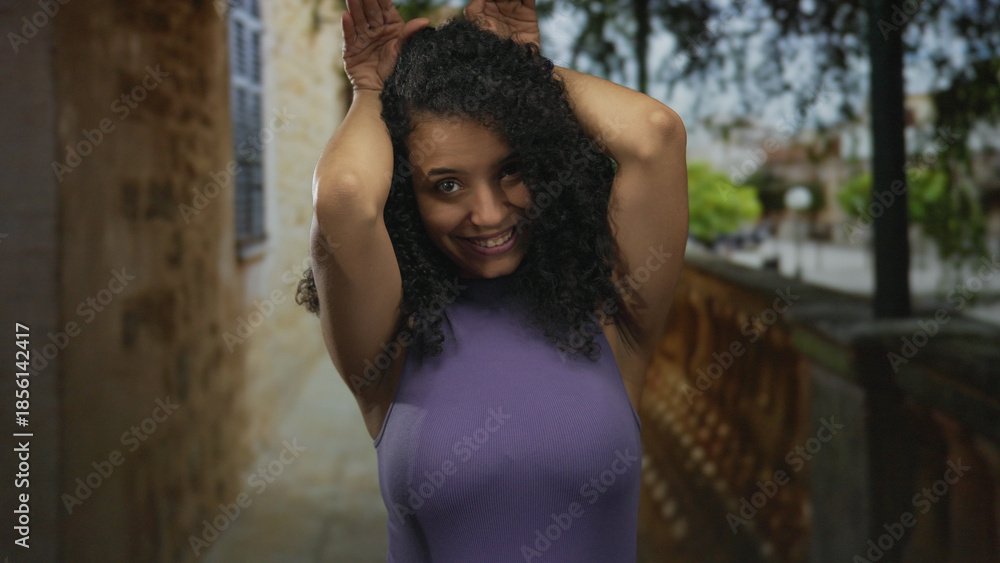 Naklejka premium Hispanic woman making ear gesture with hands on head in outdoor street walkway beside stone railing; playful moment.