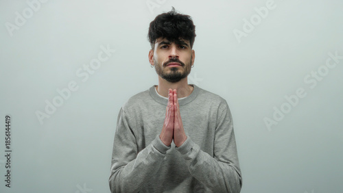 Fotografie Young man in gray sweater poses against simple white background in thoughtful prayerful gesture expressing hope and contemplation