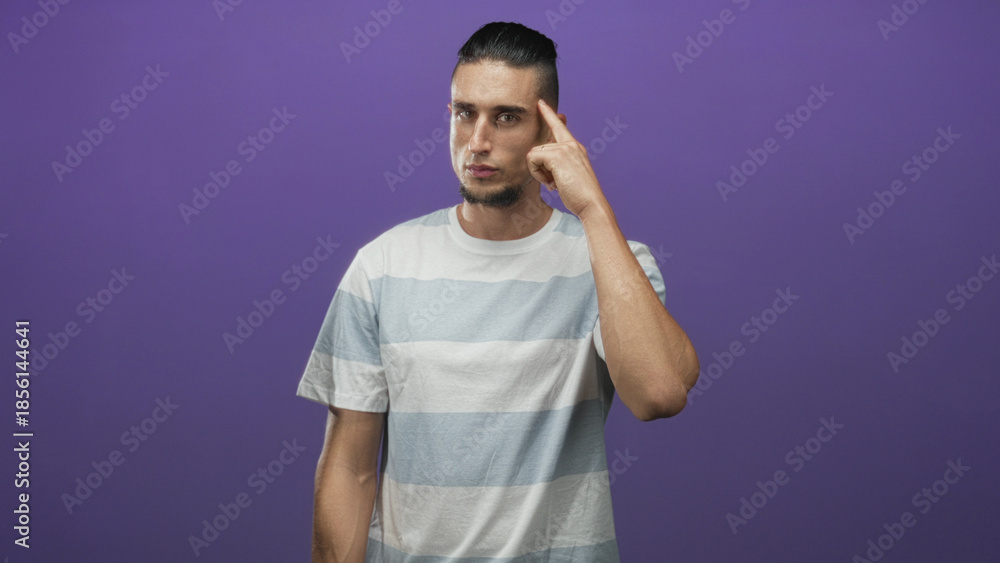 Fototapeta premium Man points finger to temple with right hand, standing in front of purple backdrop wearing striped tshirt in studio; thoughtful focus.