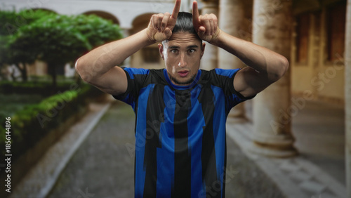 Slika na platnu Man soccer player wearing blue striped jersey points both index fingers to his head in building courtyard while squinting and leaning forward; playful taunt