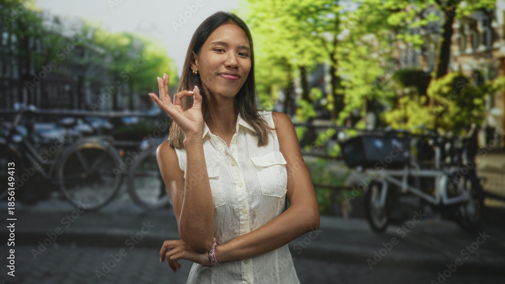 Fototapeta premium Thai woman makes ok hand gesture with visible bare hand and crossed arms on a street lined with bicycles and canal buildings; approval.