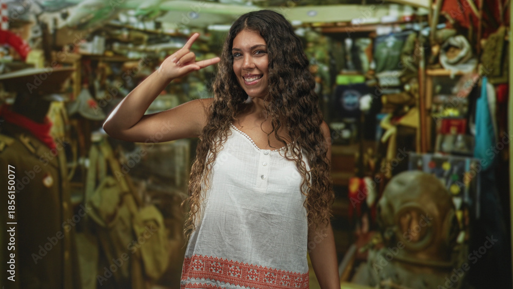 Fototapeta premium Hispanic teenage woman makes horns hand sign in an antique shop filled with vintage coats, shelves and a diving helmet; youthful joy.