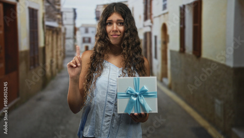 Bild auf Leinwand Woman holding gift box with blue ribbon in an old street, sleeveless top and curly hair, offers it with hesitant expression; reluctant offering uncertainty