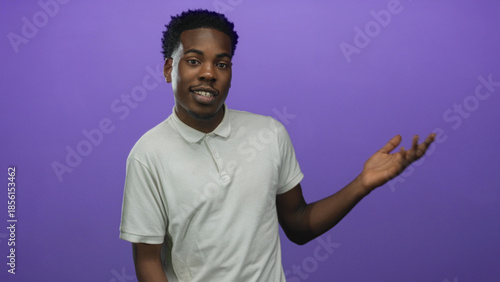 Canvas Print African american man smiling casually while gesturing open hand to side in purple studio; confidence