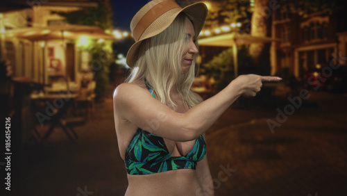 Foto Woman in straw hat pointing finger to the side on city street at night under warm lights; assertive defiance