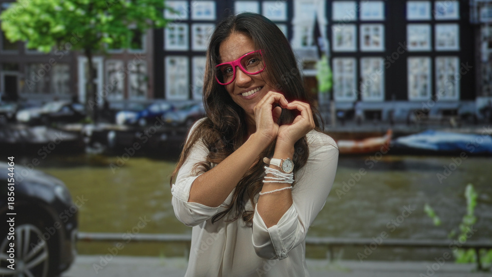 Fototapeta premium Woman wearing pink glasses forming a heart with hands on a street by an amsterdam canal, smiling; love connection.