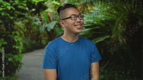 Man smiling with glasses seated on a forest path wearing a blue tshirt, looking sideways and grinning slightly; contentment.