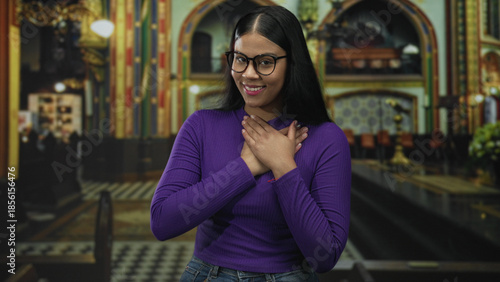 Tableau sur toile Woman with hand on chest in ornate church interior wearing glasses and purple sweater showing a thankful gesture; serenity devotion