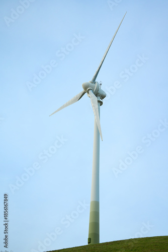 Wind turbine on hill under clear sky. Three-blade wind turbine installed on hilltop with blue sky in background.