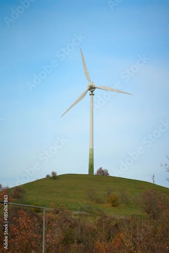 Wind turbine on hill under clear sky. Three-blade wind turbine installed on hilltop with blue sky in background.