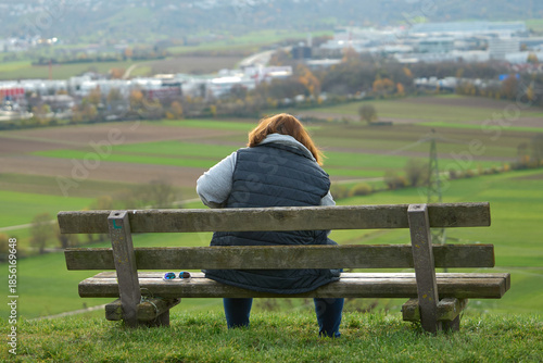 A woman sits alone on a bench. Person sits on wooden bench atop hill with city buildings and autumn trees in background.
