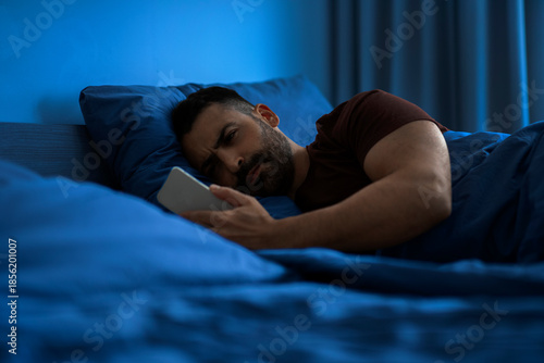 Fotografija Sleepless man lying in dark bedroom with cellphone in hand and scrolling on soci
