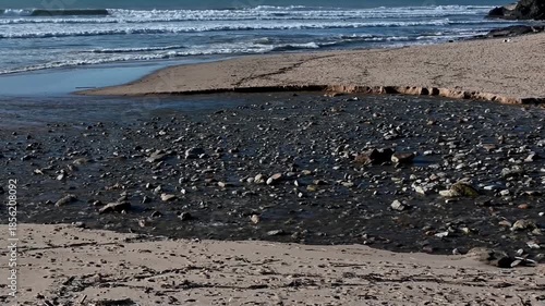 Shallow water flowing over rocks on the sand before reaching the ocean at Ericeira, Portugal. Natural coastal scene with small stream patterns, beach textures and waves in the background