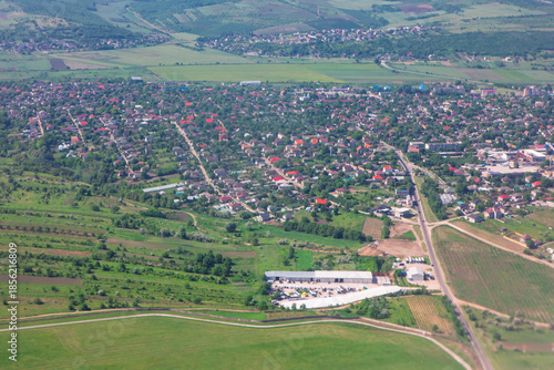 Densely populated village surrounded by vibrant green fields and rolling hills. Houses with characteristic tiled roofs line winding streets, with industrial complex visible
