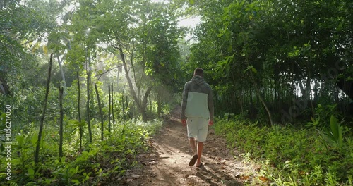 Man walks on a dirt path surrounded by trees, sunlight shining through the leaves. Tropical location. Back view. Camera follow. 