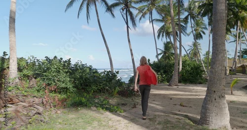 Woman exploring tropical sandy beach near the ocean, surrounded by tall palm trees.  Camera follow. 