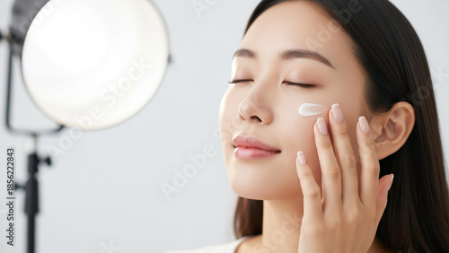 Beautiful young Asian woman gently applying moisturizing face cream to her cheek, showcasing a serene and glowing natural skincare routine in a studio setting