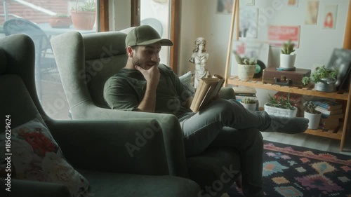 Smiling young man sitting in cozy living room, comfortably reading a book and enjoying a quiet moment of relaxation indoors.

