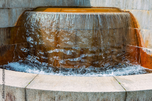 Water cascades gently over rounded tiers of stone fountain, creating smooth flow collected in lower basin. Reddish tint of stone adds earthy texture and visual warmth to tranquil architectural feature