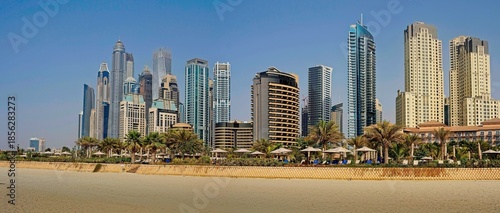 A scenic view of a white sandy beach lined with palm trees, set against a backdrop of towering modern residential skyscrapers under a blue sky.