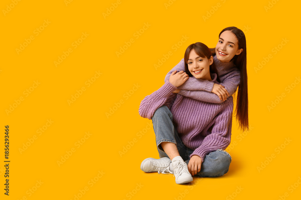 Fototapeta premium Beautiful young twin sisters hugging and sitting on yellow background