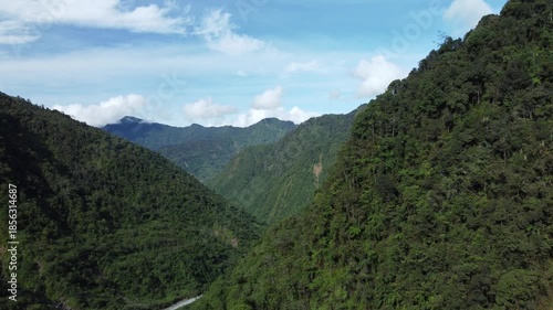 A panoramic view of a mountainous landscape with blue skies and fluffy clouds. An aerial drone moves above the dense forest canopy.