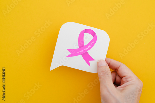 hand holding real white speech bubble paper cut with pink ribbon icon and cancer awareness text on yellow background