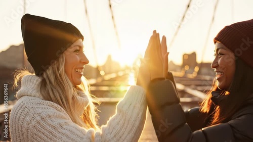Two happy young women in knit hats high fiving on a bridge during sunset with a city skyline