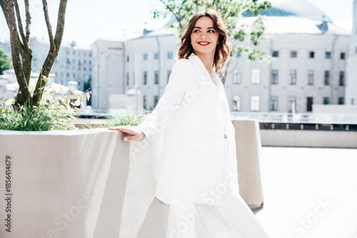 Young beautiful brunette woman wearing nice trendy white suit jacket. Sexy smiling model posing in the street at sunny day. Fashionable female outdoors. Cheerful and happy. Evening makeup, red lips